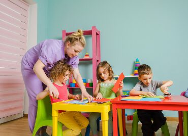 Teacher Helping Children With Crafts At A Colorful Classroom Table